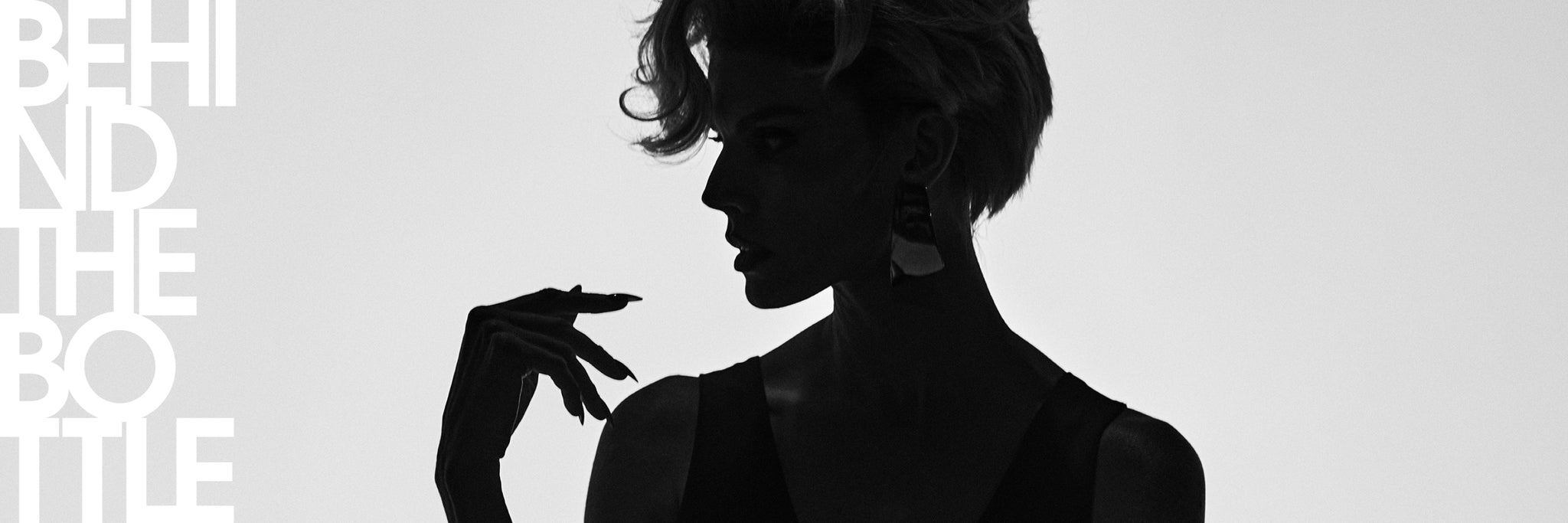 A high-contrast black and white editorial photograph of a woman in profile, silhouetted against a light background, with large white text on the left reading 'BEHIND THE BOTTLE'.